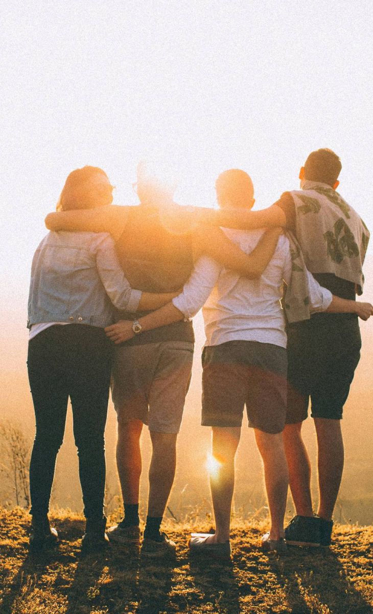Four friends stand arm-in-arm on a hill at sunset, looking out over a scenic landscape.