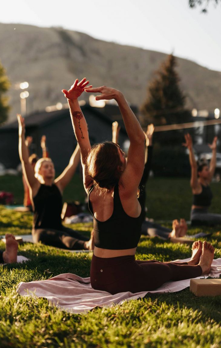 People practicing yoga outdoors on grass, stretching arms upward, with mountains in the background.