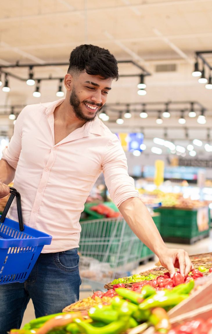 Smiling man with a basket picks fresh produce in the vegetable section of a grocery store.