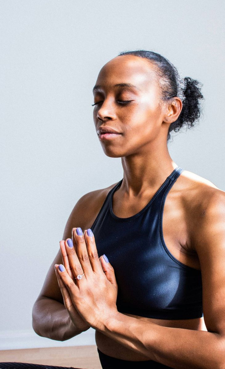 Woman in workout clothes sitting cross-legged, eyes closed, hands in prayer position, meditating indoors.